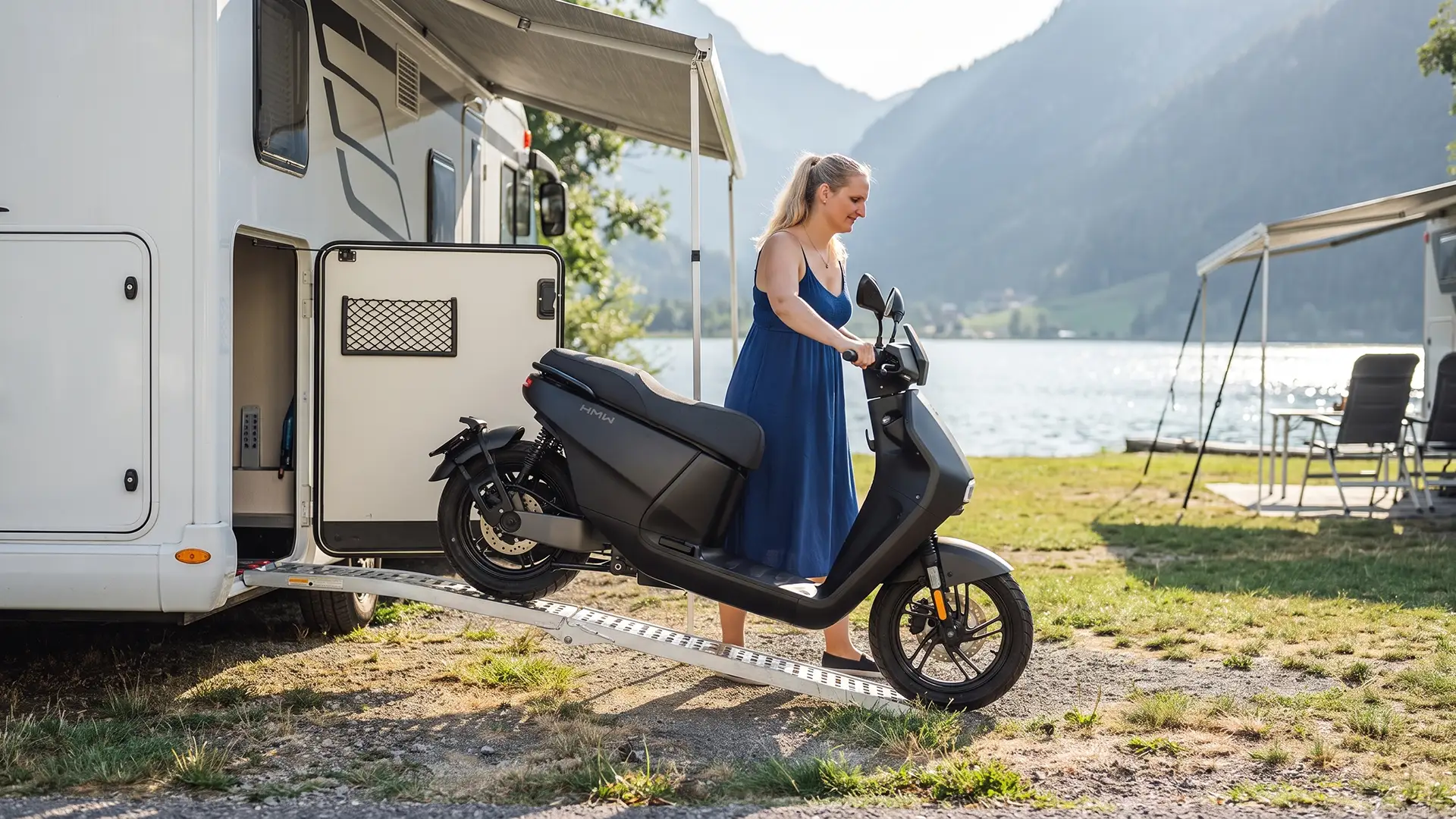 Eine junge Frau lädt eine HMW ConnE aus dem Stauraum eines Campingvans, der auf einem Campingplatz eines Alpensees geparkt ist.